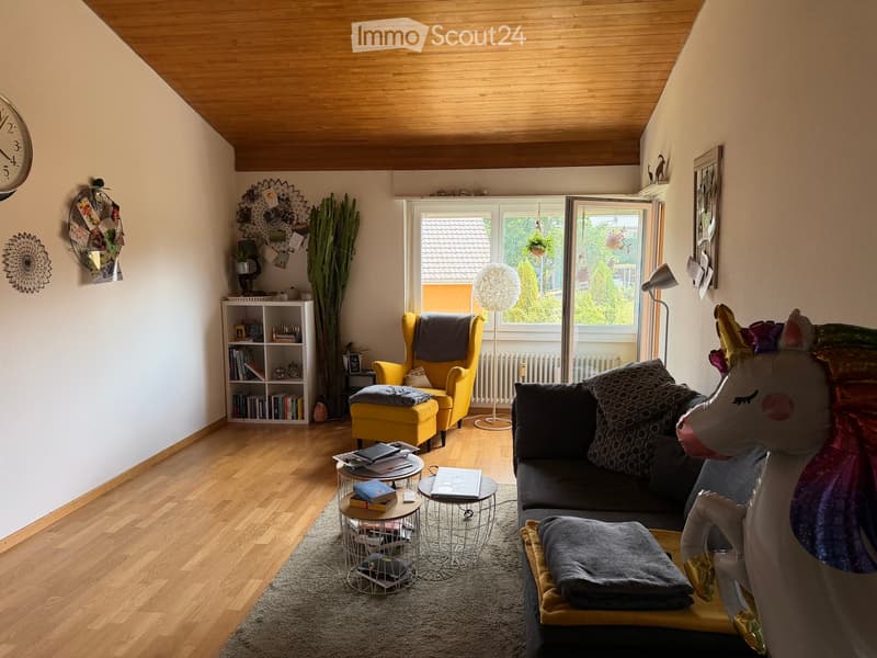 Living room with wooden flooring, a grey sofa, yellow armchair, small round tables, books, a shelf, and a decorative unicorn toy.