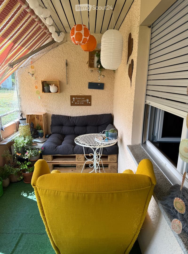 Cozy balcony with a green carpet, yellow armchair, dark grey sofa, small white table, and several potted plants.