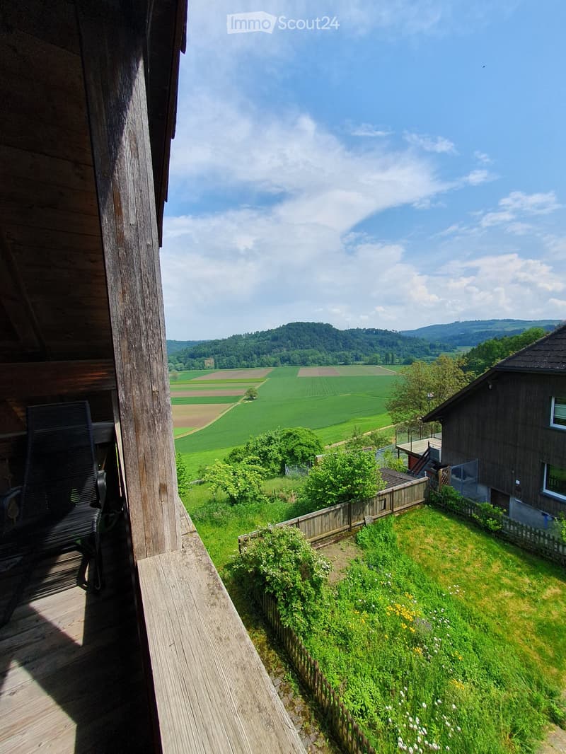 Balcony with wooden decking, overlooking a green valley with fields and hills under a partly cloudy sky. A single chair is placed on the balcony.