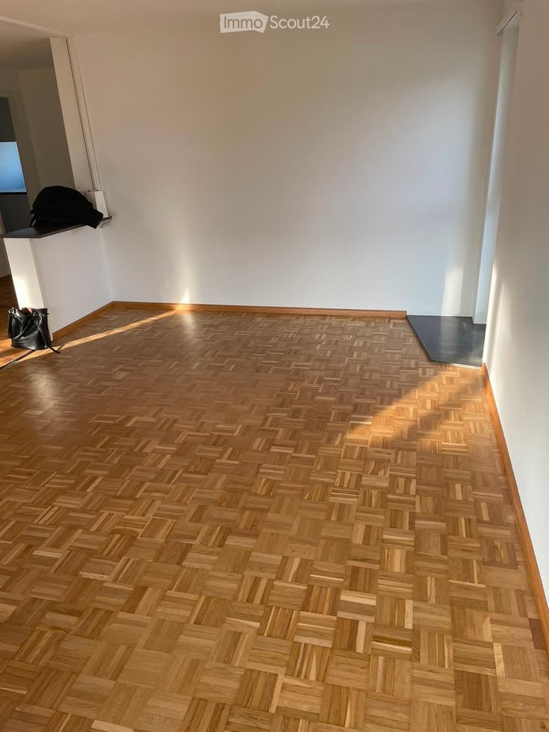 Empty room with wooden herringbone flooring and white walls, featuring a small shelf with a black bag and a black mat near the doorway.