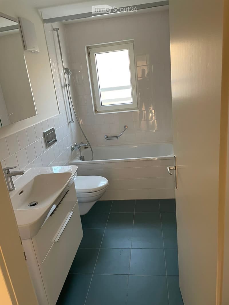Modern bathroom with a sink, toilet, bathtub, and a window above the tub. Features a dark tiled floor and light-colored walls.