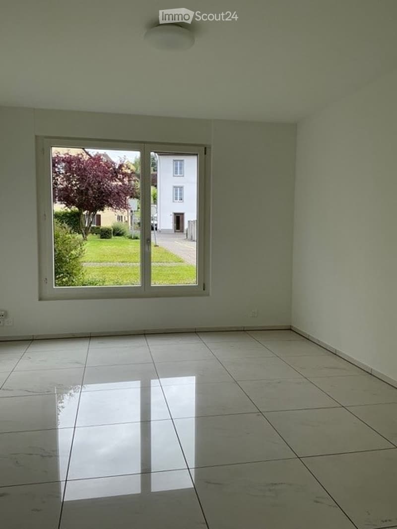 Empty room with tiled floor, large window showing a view of a garden and neighboring house, white walls, and a ceiling light fixture.