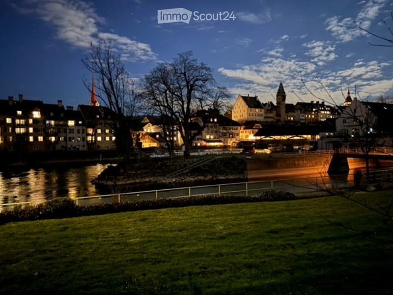 Nighttime view of a town with illuminated buildings, a river, and a park area in the foreground.