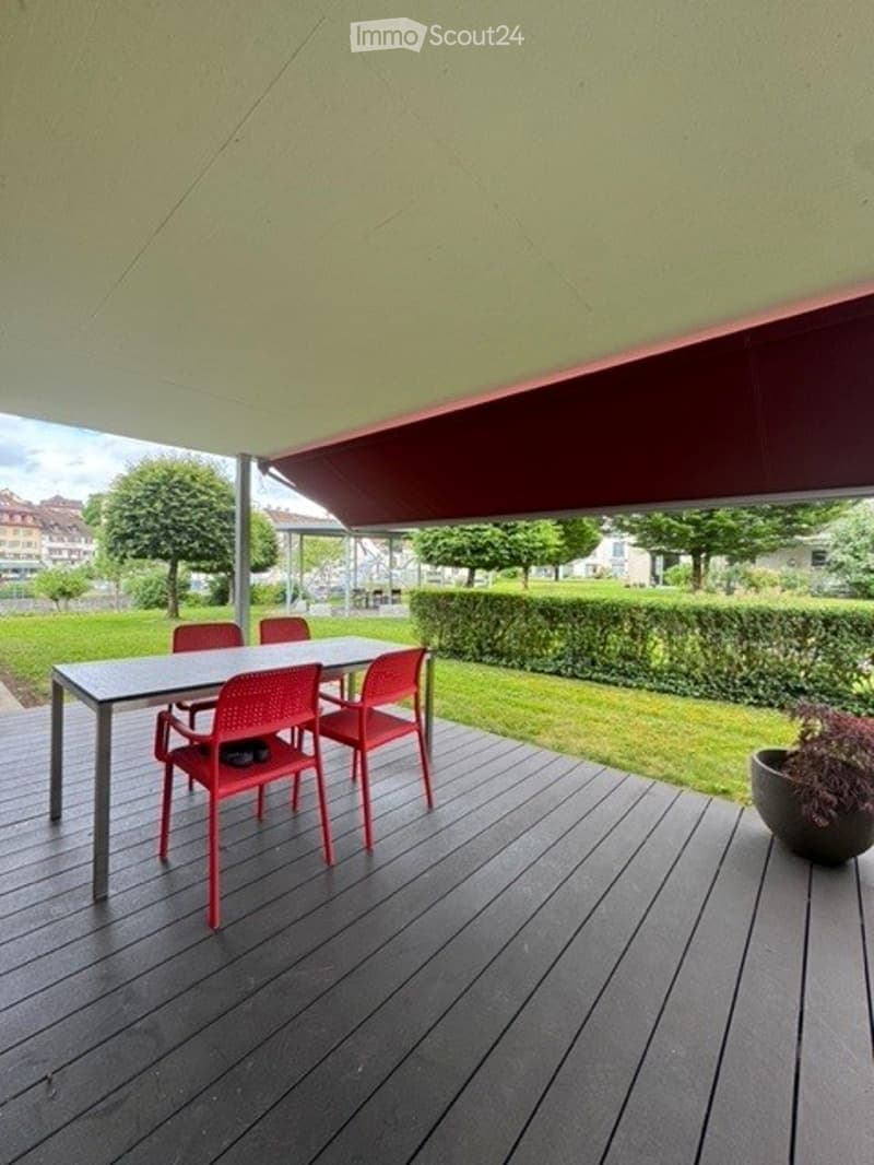 A wooden deck with a table and red chairs, covered by a large awning, surrounded by a well-maintained lawn and trees.