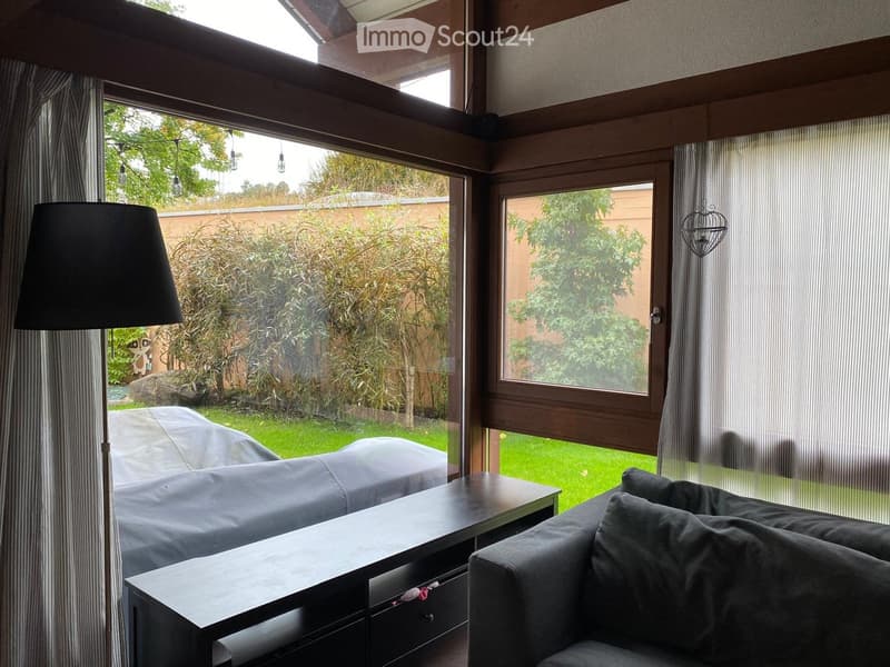 Modern living room with large windows, grey sofa, coffee table, and floor lamp. The room features wooden beams on the ceiling and striped curtains.