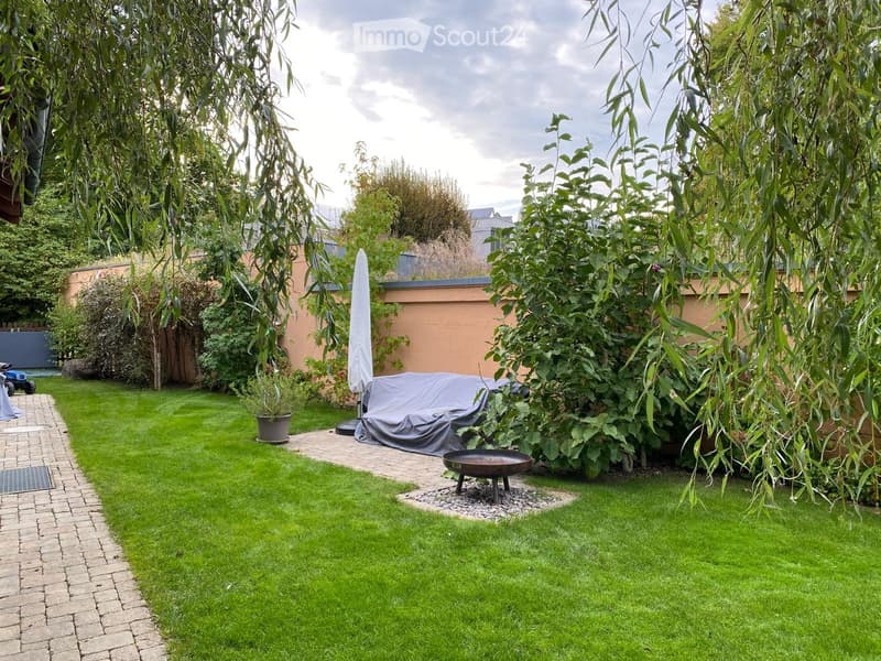 Well-maintained garden with a neatly trimmed lawn, brick pathway, potted plants, a covered outdoor seating area, and a fire pit.