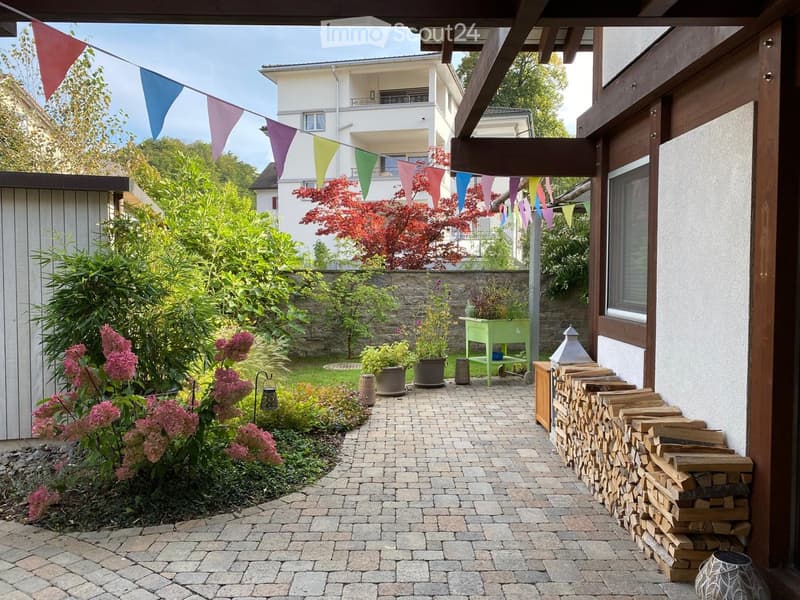 Garden area with a paved pathway, potted plants, a stack of firewood, and colorful triangular flags strung overhead.