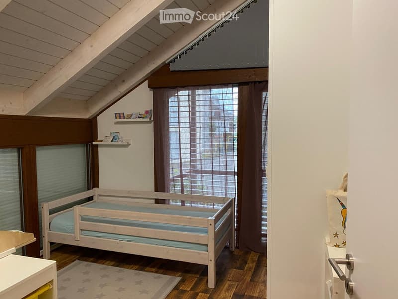 Cozy attic bedroom with wooden slanted ceiling, wooden bed with blue mattress, small shelf with picture frames, wooden chair, and a star-patterned rug.