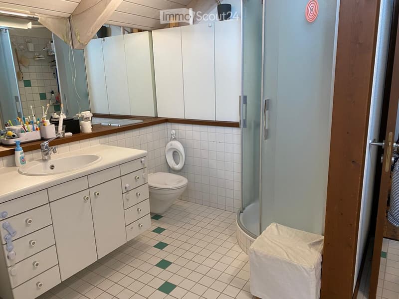 Bathroom with white tiles, a sink with a faucet, a toilet, a shower, and a mirrored cabinet. The countertop holds various toiletries including toothbrushes and toothpaste.