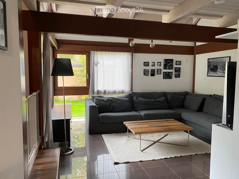 Modern living room with a grey sectional sofa, wooden coffee table, floor lamp, and large windows with sheer curtains. The room features a mix of dark and light tones, with picture frames on the wall and a view of a green garden outside.