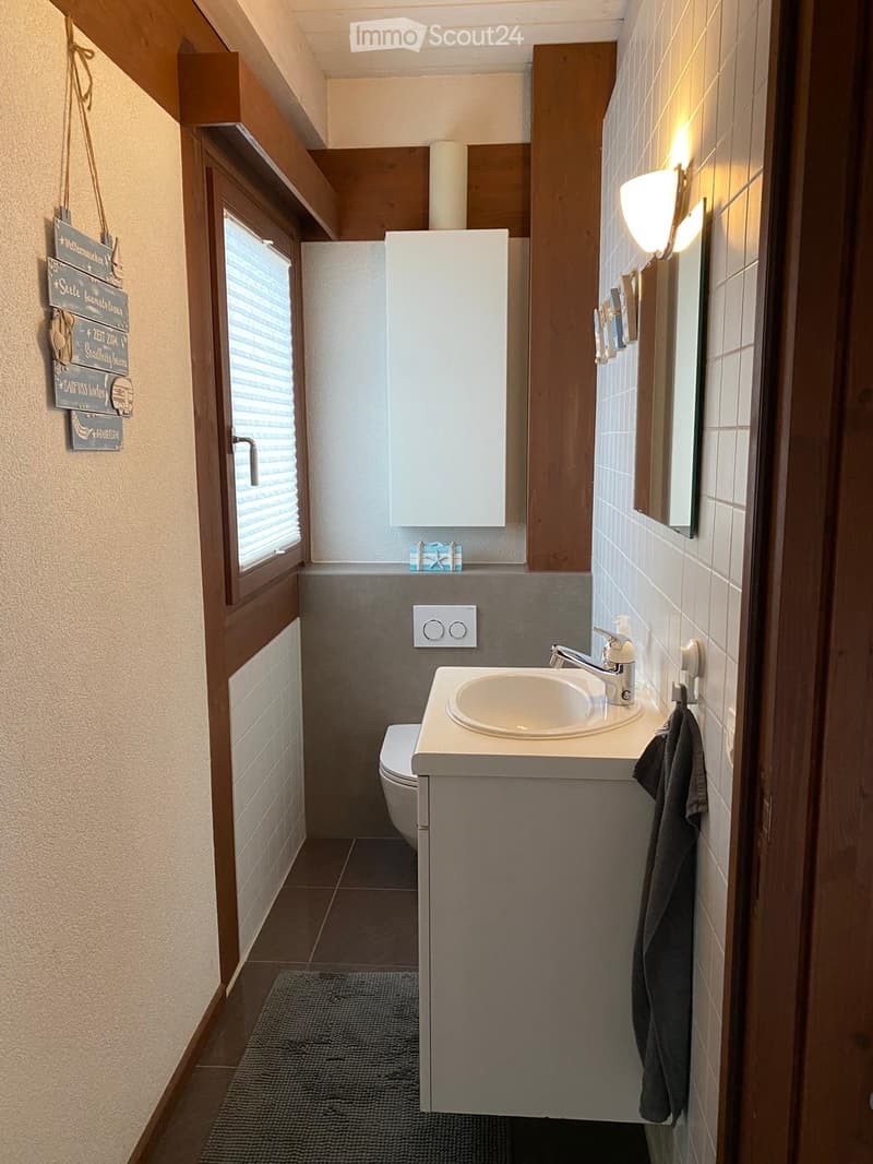 Modern bathroom with a white sink, chrome faucet, mirror, toilet, and a towel hanging on the wall. The room features wooden trim, tiled walls, and a small window with blinds.