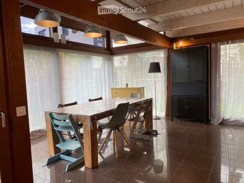 Bright dining area with a large wooden dining table, multiple chairs, a tall floor lamp, and a dark cabinet. Large windows with sheer curtains allow natural light to fill the space.