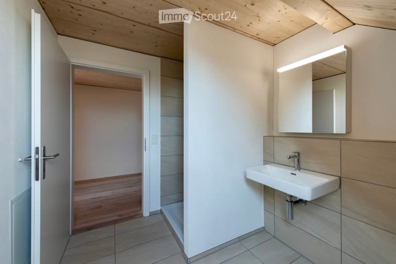 Modern bathroom with a white sink, mirror, and wooden flooring. The room features a sleek design with a built-in mirror and a door leading to another space.