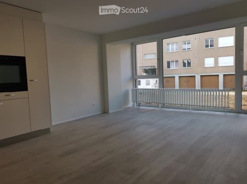 Empty room with wooden flooring, large windows, and a balcony railing visible through the windows.