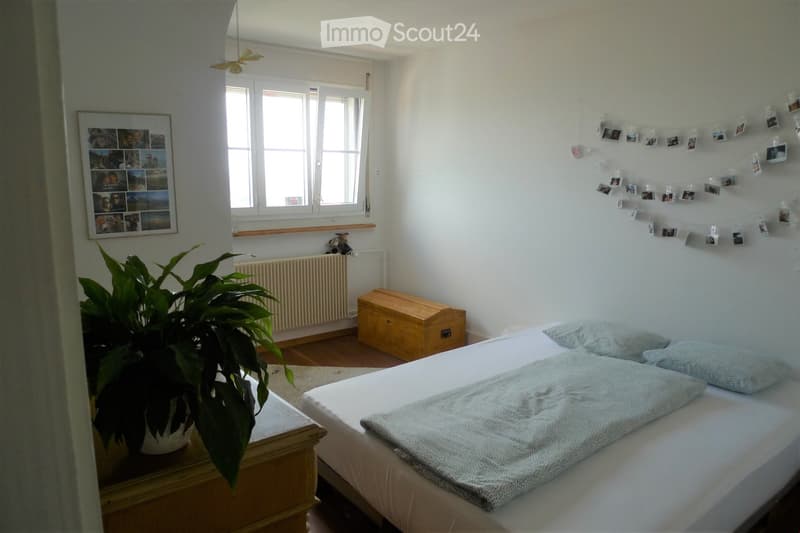 Cozy bedroom with a double bed, nightstand, plant, radiator, wooden chest, picture frame, and photo string on the wall.