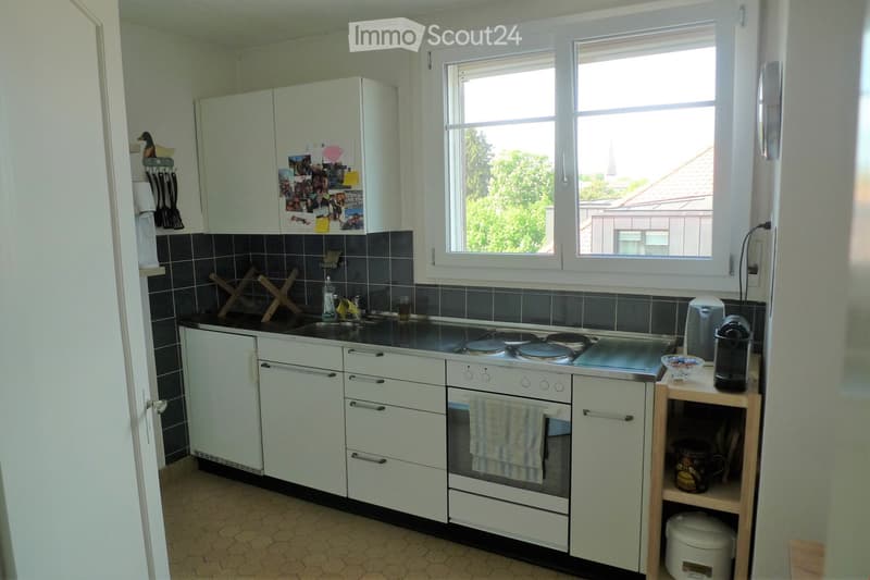 Bright kitchen with white cabinets, a black countertop, and a built-in oven. The space features a sink with a faucet, a dish rack, and various appliances including a coffee maker and a small wooden shelf with cups and a bowl. A towel hangs from the oven h