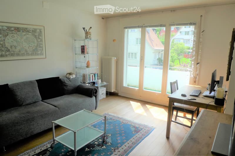 A modern living room with a grey sofa, glass coffee table, wooden floor, large windows, a desk with a laptop, and a bookshelf with books and decorative items.