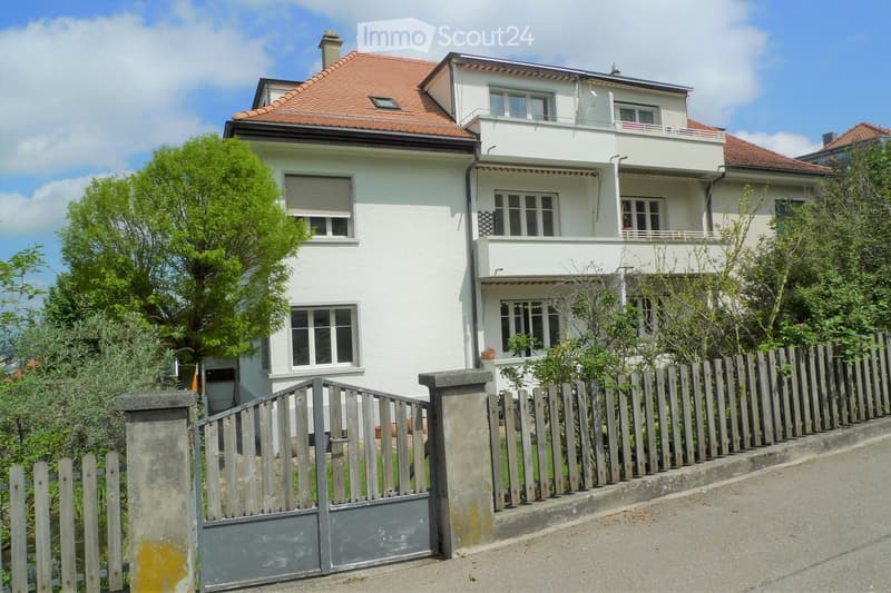 Two-story white house with red-tiled roof, multiple windows, and a balcony on the upper floor. The house is surrounded by a wooden fence and greenery.