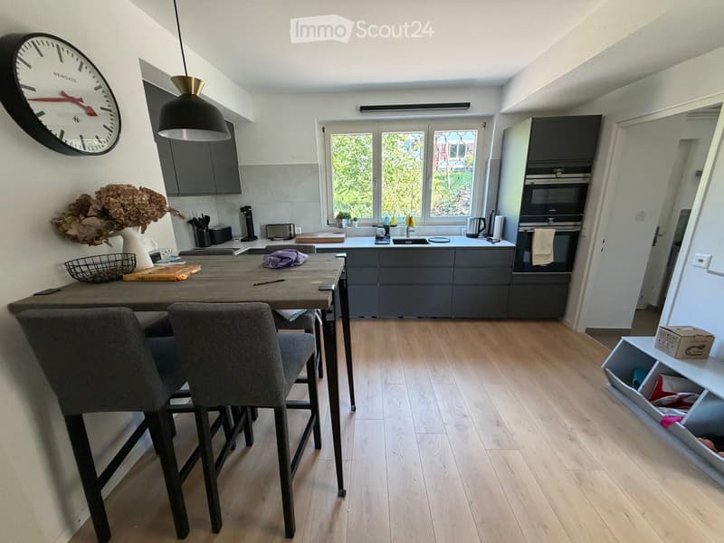 Modern kitchen with grey cabinets, wooden floor, central island, dining table with chairs, and various appliances including an oven, sink, and coffee maker.
