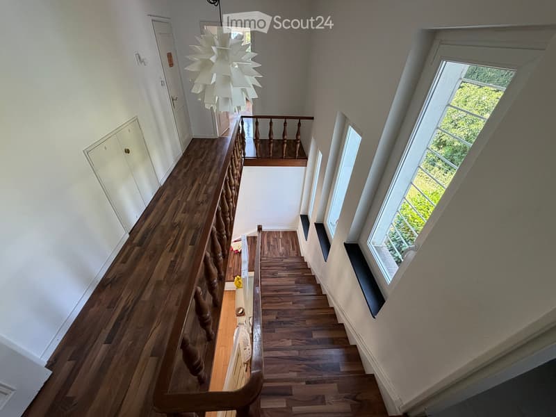 Spacious hallway with wooden flooring, white walls, and a staircase leading to an upper level. Features a decorative chandelier and large windows allowing natural light to enter.