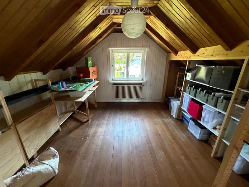 Spacious attic room with wooden floors, slanted wooden ceilings, large window, shelves, and storage boxes.