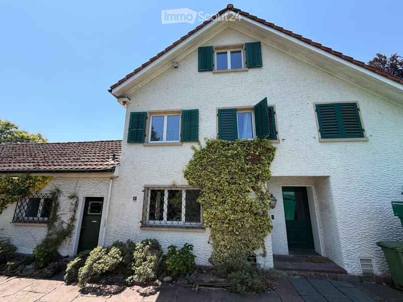 Two-story white house with green shutters, ivy-covered walls, and a tiled roof. Features include a front door, multiple windows, and a small garden area with shrubs.