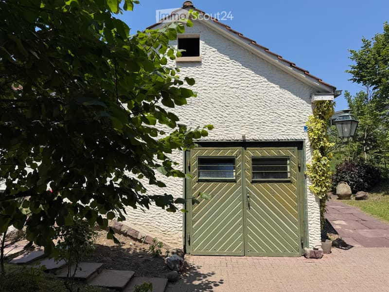 White stone building with a green garage door, surrounded by greenery and a paved pathway.