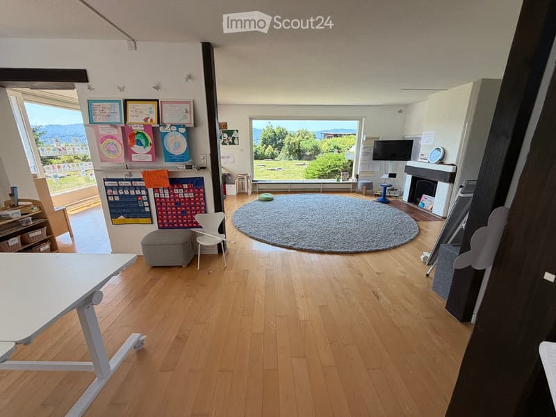 Bright room with wooden flooring, large windows, round rug, table, chairs, calendar, and fireplace.