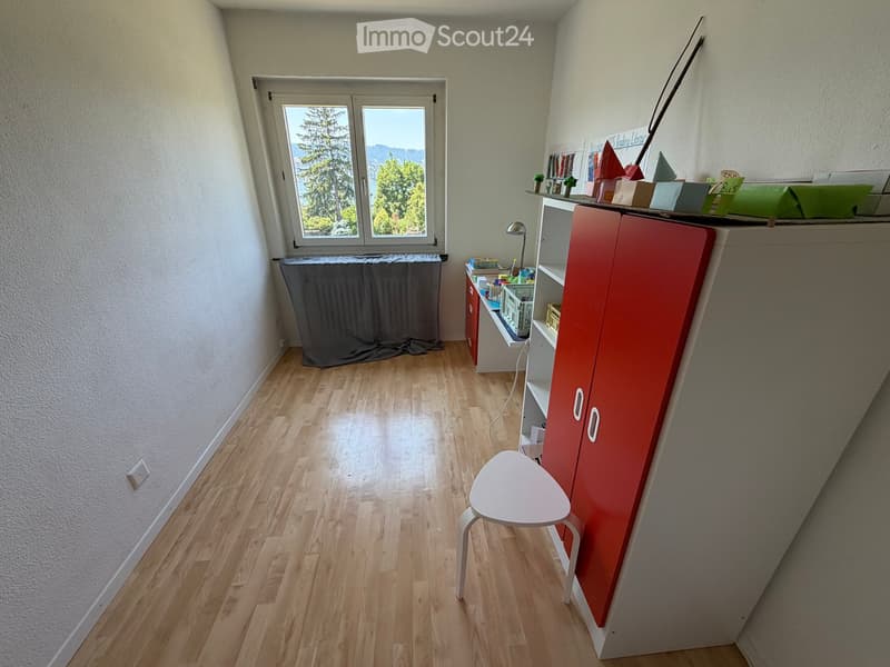 A small, well-lit room with wooden flooring, a large window offering a scenic view, a red cabinet, a white stool, and a small shelf with books and decorative items.
