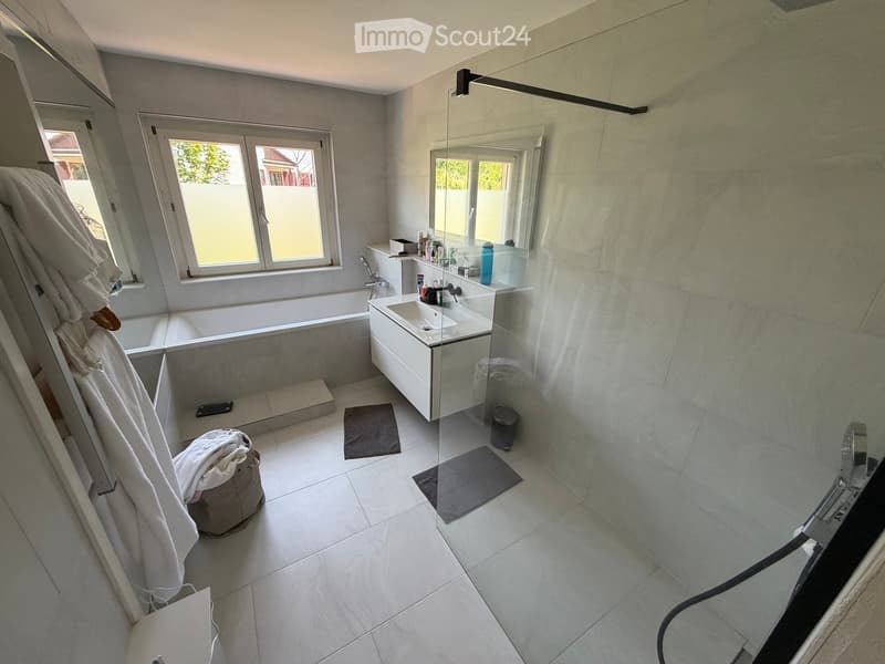 Modern bathroom with a bathtub, sink, mirror, and various toiletries. Features tiled walls and floor, a window, and towels on the side.