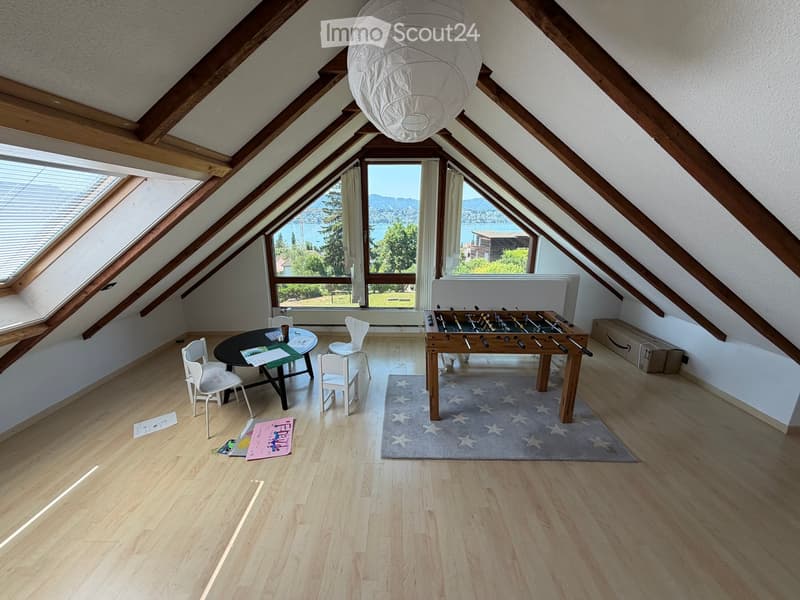 Bright attic room with wooden beams, large windows offering scenic views, a round table with chairs, and a football table.