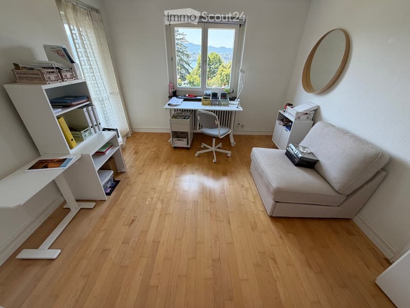 A cozy, well-lit room with wooden flooring, a sofa, a desk with a chair, a shelf, and a mirror on the wall. The room features large windows with a scenic view.