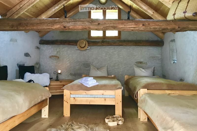 Cozy attic bedroom with wooden beams, three wooden beds with white and brown bedding, a nightstand with a lamp, a hat on a shelf, a bench, and a fur rug on the floor.