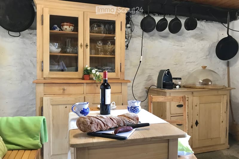 A rustic kitchen with wooden cabinets, a central island, and a dining table set with bread, a bottle of wine, and cups. The cabinets are filled with glassware and dishes, and a coffee maker sits on the island.