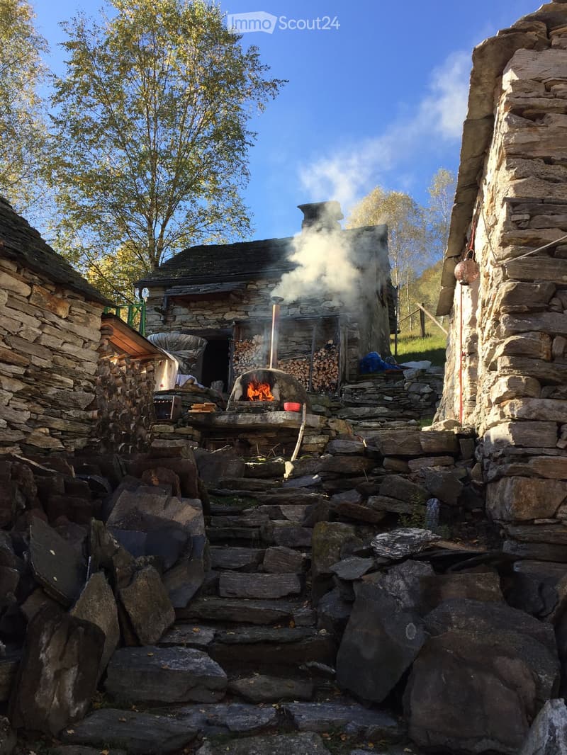 Stone outdoor fireplace with wood stacked nearby, surrounded by stone walls and steps, with smoke rising from the fire.
