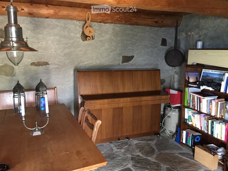 A rustic dining area with a wooden table, chairs, a bookcase filled with books, and a piano. The room features exposed wooden beams, stone walls, and a hanging lantern.