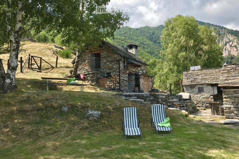 A rustic stone cottage surrounded by a grassy area with two striped lounge chairs, a picnic table, and a stack of firewood. The setting is surrounded by trees and hills.