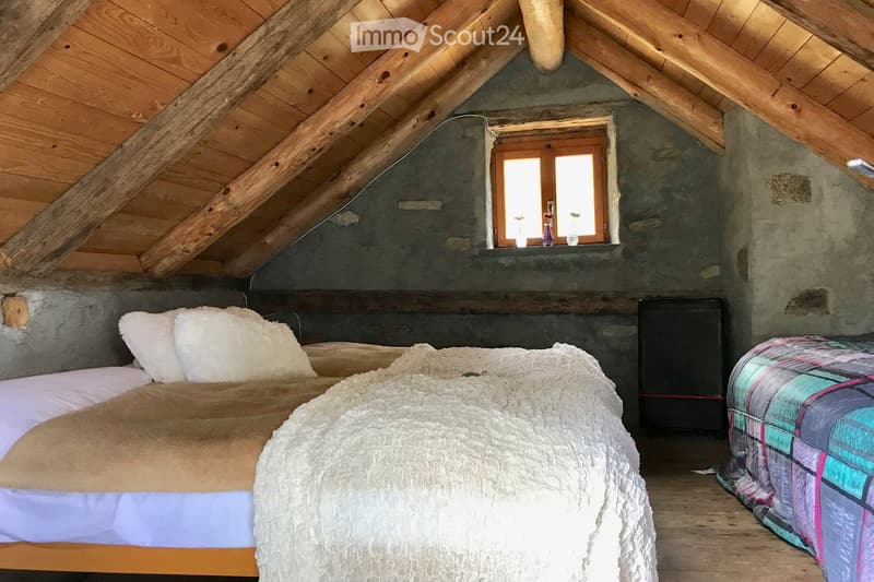 Cozy attic bedroom with wooden beams, exposed wooden ceiling, and a large window. Features a double bed with white pillows and a fluffy white blanket, a nightstand with a vase, and a colorful quilt on a second bed.