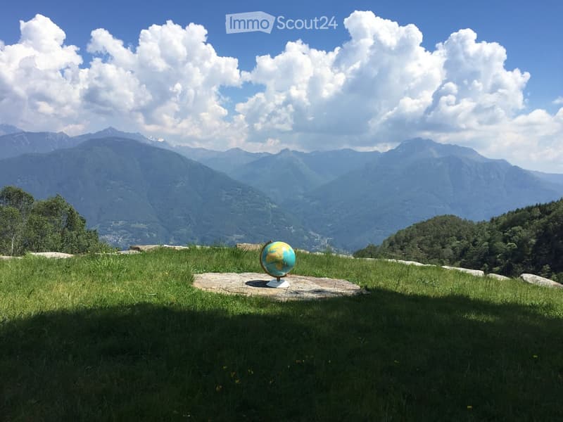 A grassy hilltop with a globe on a stone platform, overlooking a mountain range under a partly cloudy sky.