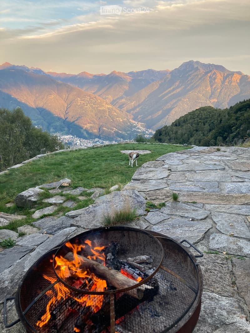 A scenic outdoor area with a stone fire pit burning, surrounded by rocks and grass, with a dog nearby and a panoramic mountain view in the background.