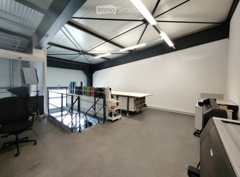 Open-plan room with exposed metal beams and skylights, featuring a large worktable, shelves with supplies, a chair, and a printer.