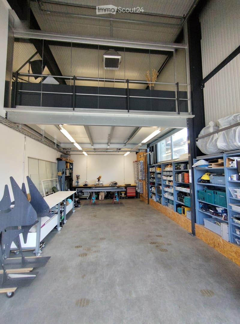The image shows a spacious, industrial-style workshop or studio with concrete floors, exposed metal beams, and large windows. Visible objects include tables, shelves, stools, and cabinets.