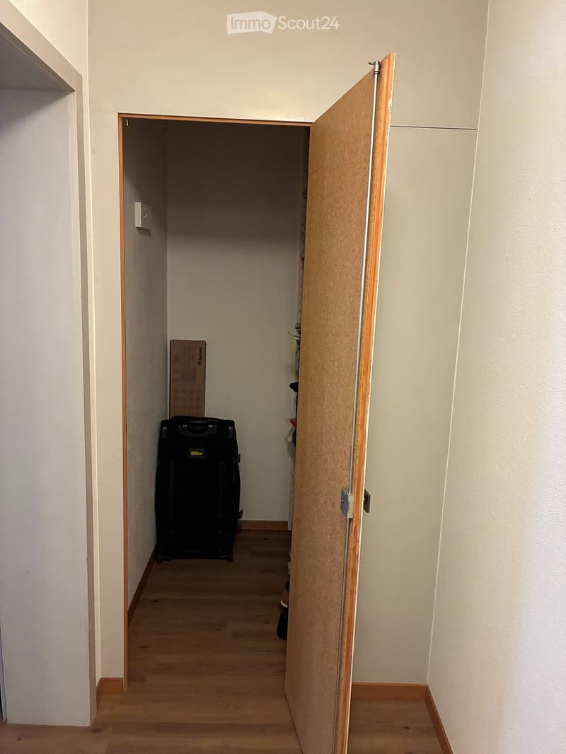 A narrow hallway with light wooden flooring and white walls, featuring an open door leading to another room where a suitcase is visible.