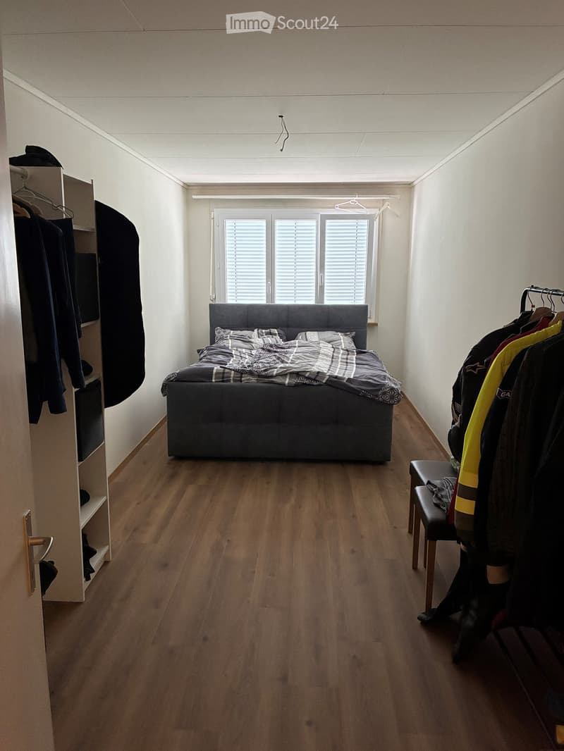A modern bedroom with a dark grey upholstered bed, patterned bedding, a small wooden stool, and a built-in wardrobe with hanging clothes.