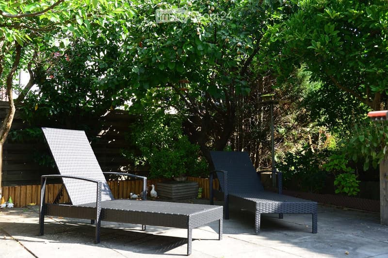 A sunny outdoor patio with two lounge chairs, a small table, and lush greenery, including fruit trees and decorative plants.