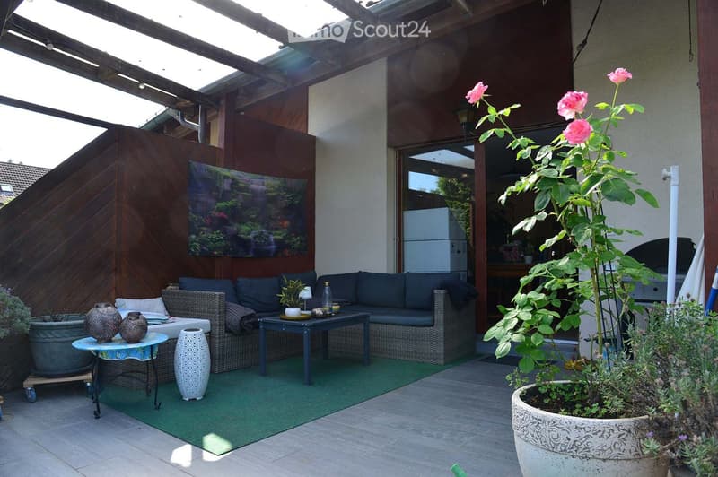 Outdoor patio with a green artificial turf rug, wicker sofa, small table, decorative vases, and potted plants, including a large rose bush and a small tree.