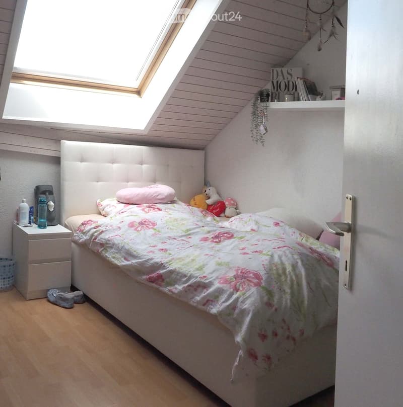 Cozy bedroom with a white upholstered headboard, floral duvet, nightstand with bottles and a cup, and decorative toys on the bed.