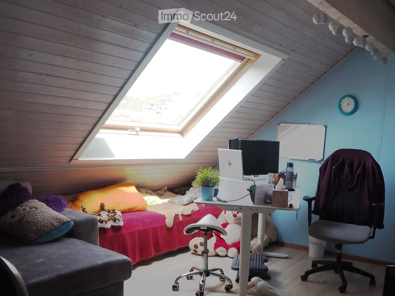 Cozy attic room with a bed, desk, laptop, chair, and decorative items. Features a skylight window and a small plant on the desk.