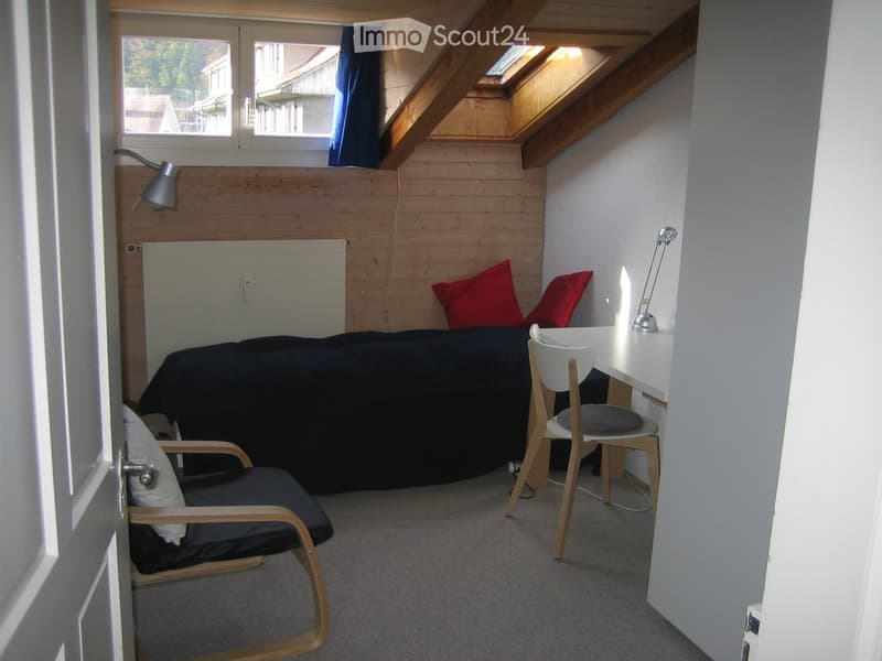 Cozy attic bedroom with wooden walls, slanted ceiling, bed, desk, chair, and two lamps.