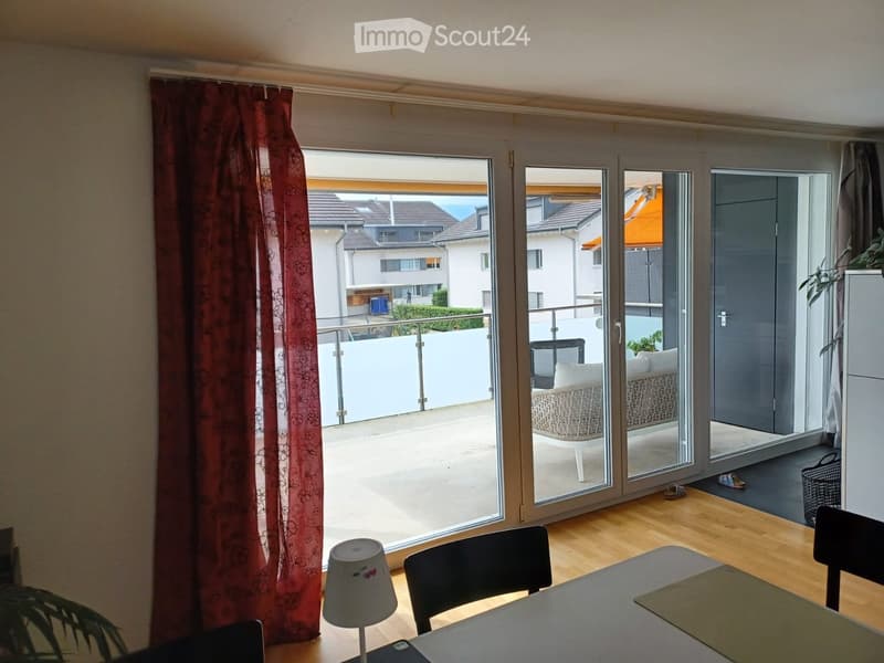 Bright room with wooden flooring, large sliding glass doors leading to a balcony, a small table with chairs, and a lamp on a side table.